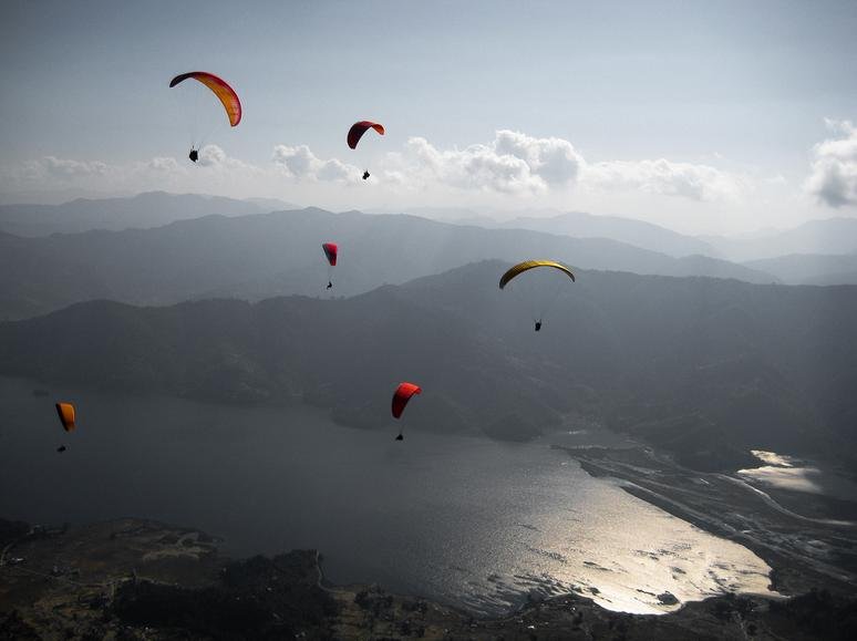 Six colorful paragliders flying over Phewa Lake in Pokhara, Nepal, with mountains in the background.