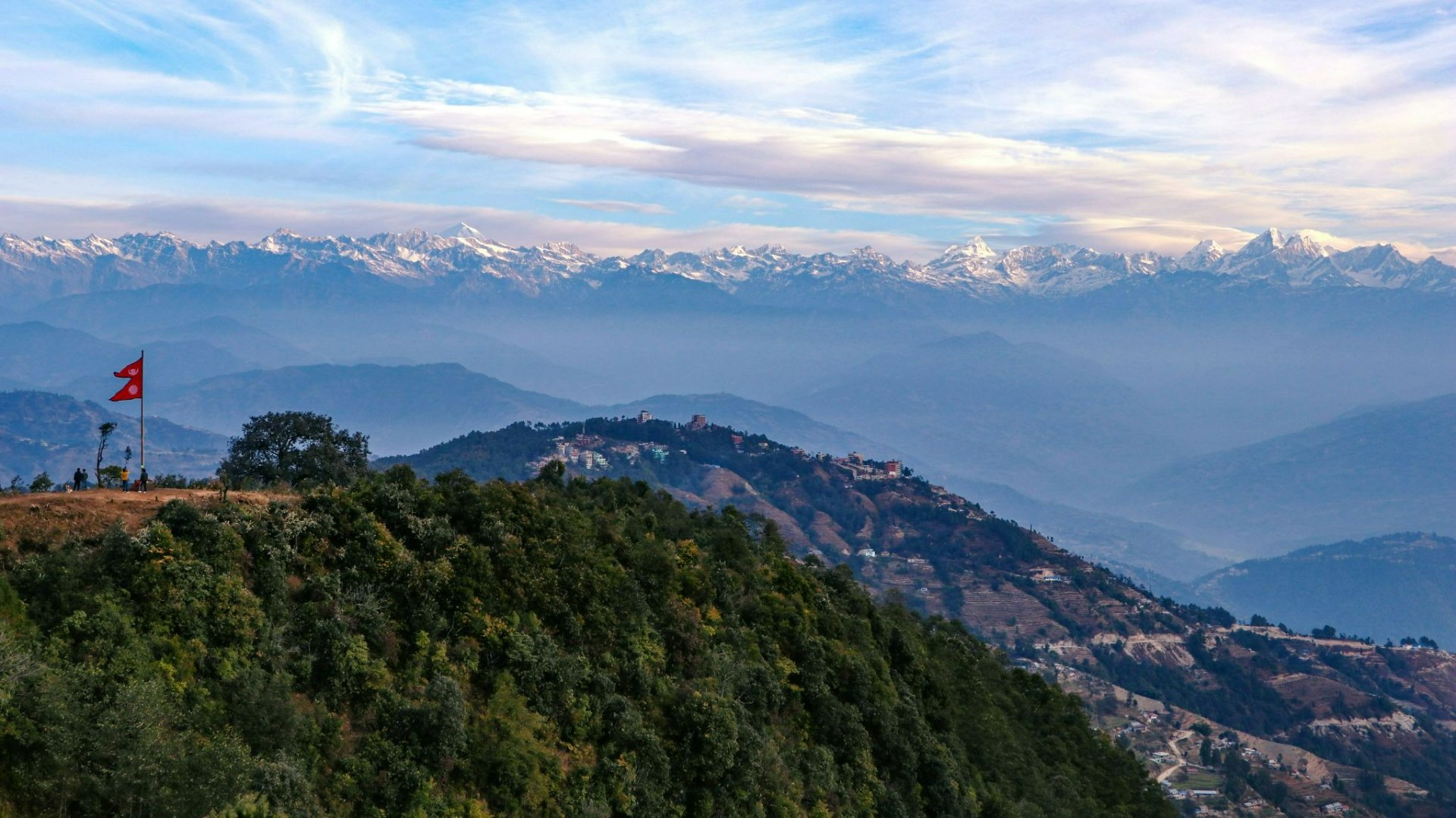 Nepal flag on open ridge viewpoint with panoramic Himalayan range and Nagarkot village below