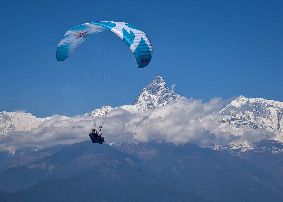 A paraglider with a green canopy flying in front of the snow-covered Machhapuchhre (Fishtail Mountain) in Pokhara, Nepal, under a clear blue sky.